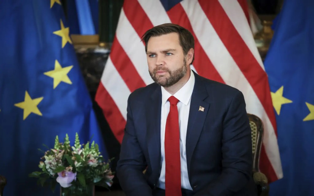 A man sitting attentively in front of American and European flags, with a bouquet of flowers in the foreground.