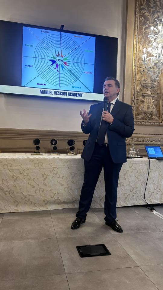 A speaker at the Manuel Vescovi Academy presents during the 'Tuscany in the World' award ceremony, with a projection of a colorful diagram in the background.