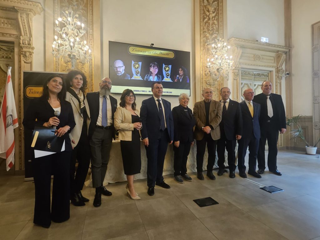 Group photo of award recipients and presenters at the 'Tuscany in the World' ceremony in the historic Palazzo della Regione, Florence, featuring a backdrop with event details.