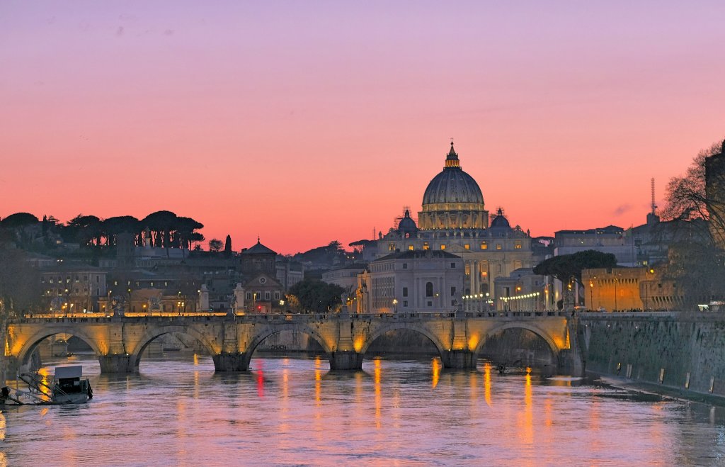 A picturesque view of the Vatican at sunset, showcasing St. Peter's Basilica and the surrounding architecture, with a vibrant pink and purple sky reflected in the river.