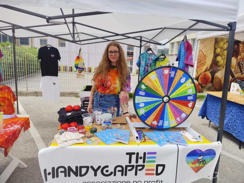 A young woman with long hair stands behind a booth at an outdoor event, displaying colorful merchandise and a prize wheel. The booth is branded with the name 'THE HANDYCAPPED' and features various items related to the project.