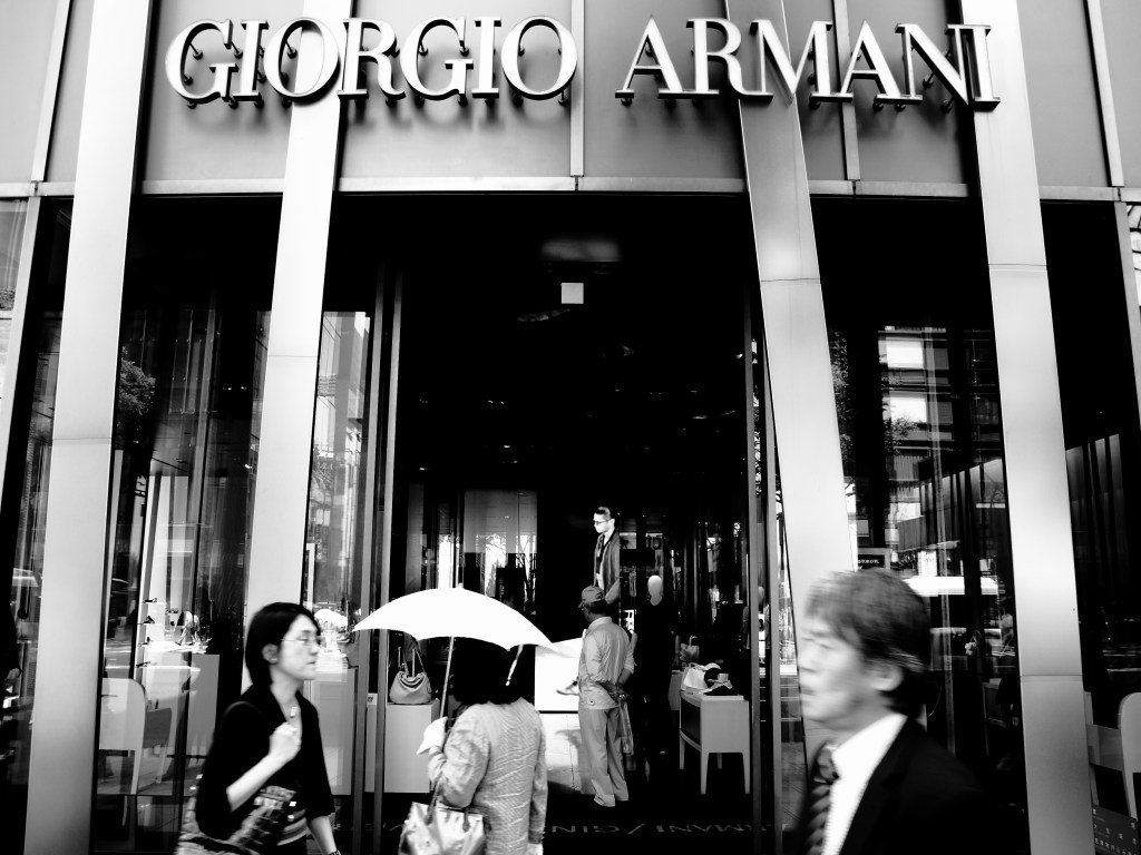Black and white image of the Giorgio Armani storefront with shoppers passing by, one holding an umbrella, showcasing the brand's elegant entrance and modern architecture.
