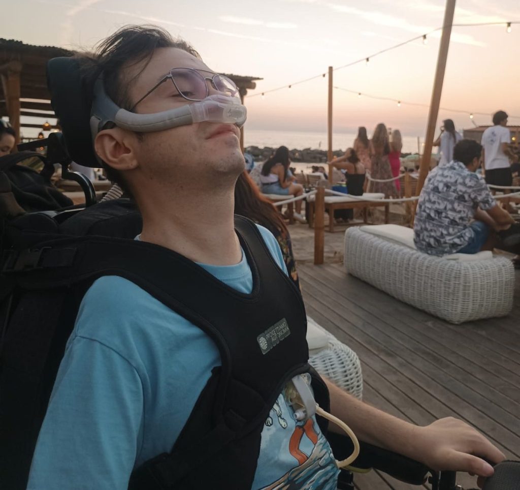 A young person in a wheelchair enjoying a sunset at a social gathering, wearing glasses and looking relaxed.
