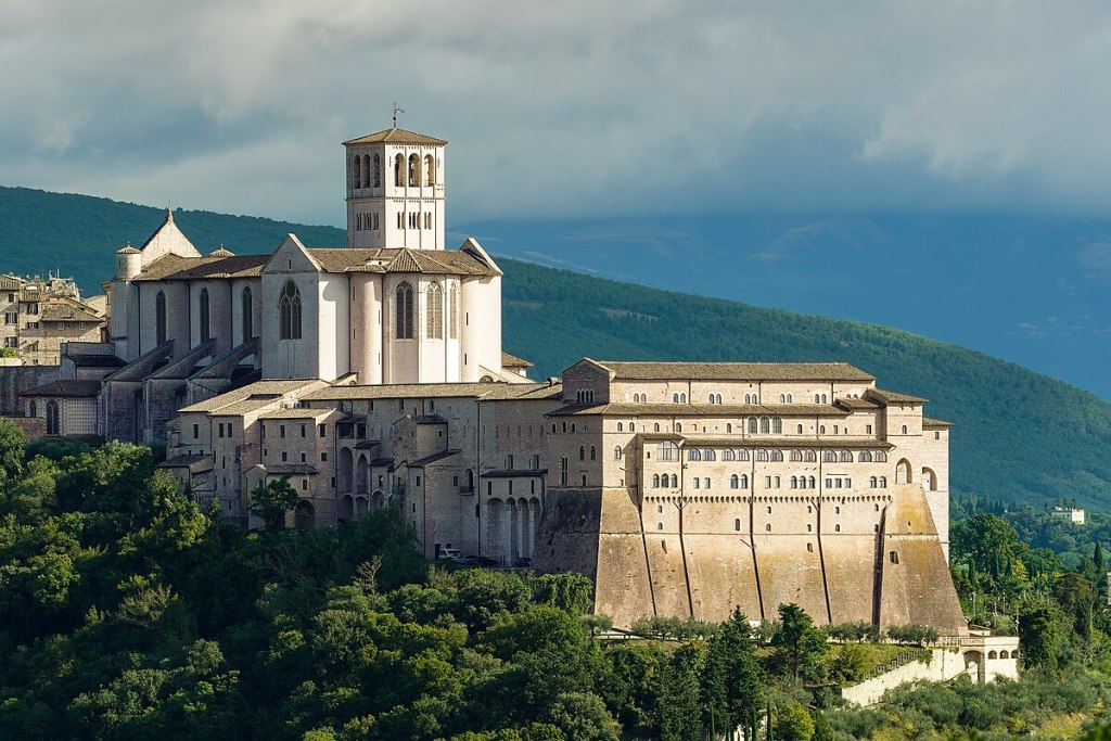 A panoramic view of the Basilica of Saint Francis in Assisi, Italy, showcasing its architectural beauty against a backdrop of greenery and hills under a cloudy sky.
