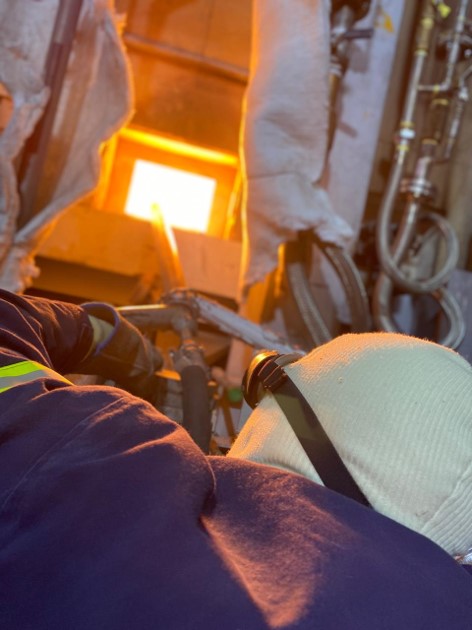 The welding lance extends deep into the glowing yellow interior of the melting furnace to reach distant damaged areas.