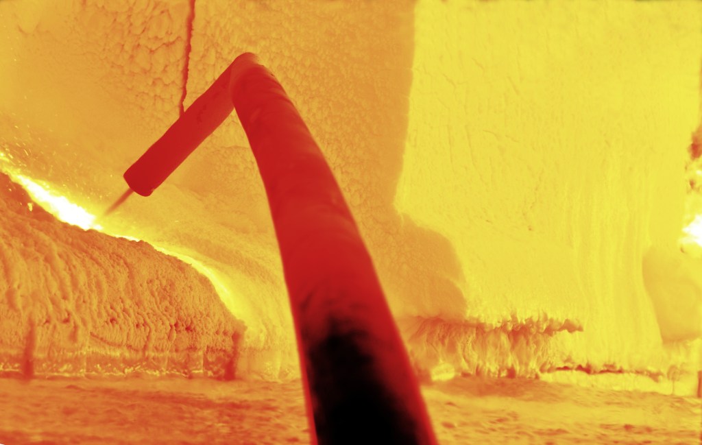 A close-up of a high-temperature industrial welding tool being used in a molten glass environment, glowing with intense orange and yellow hues.