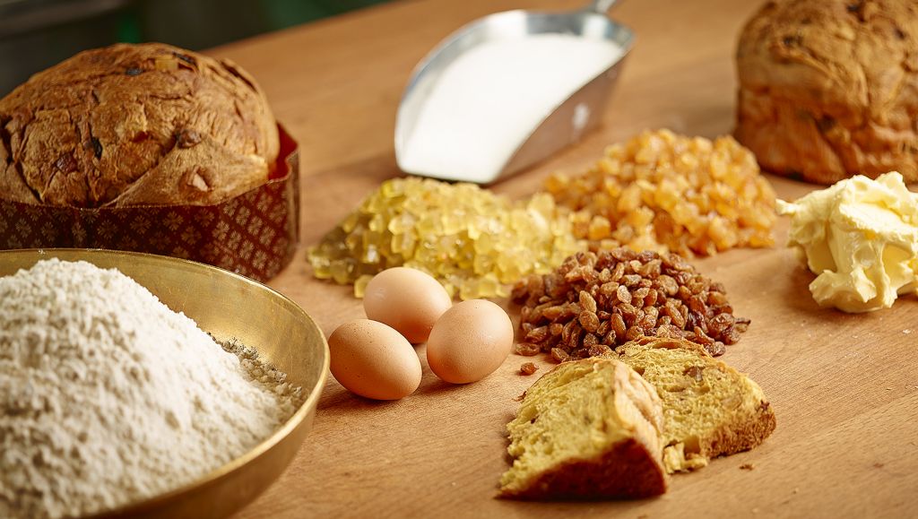 Ingredients for traditional Panettone, including flour, eggs, candied fruits, butter, and two dome-shaped Panettone loaves on a wooden surface.