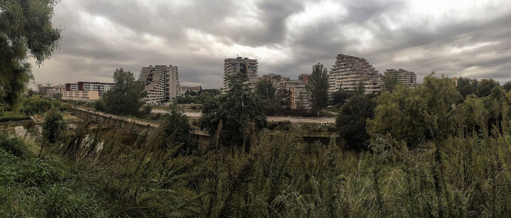 A panoramic view of a desolate urban landscape featuring modernist architecture amidst overgrown vegetation and a cloudy sky.