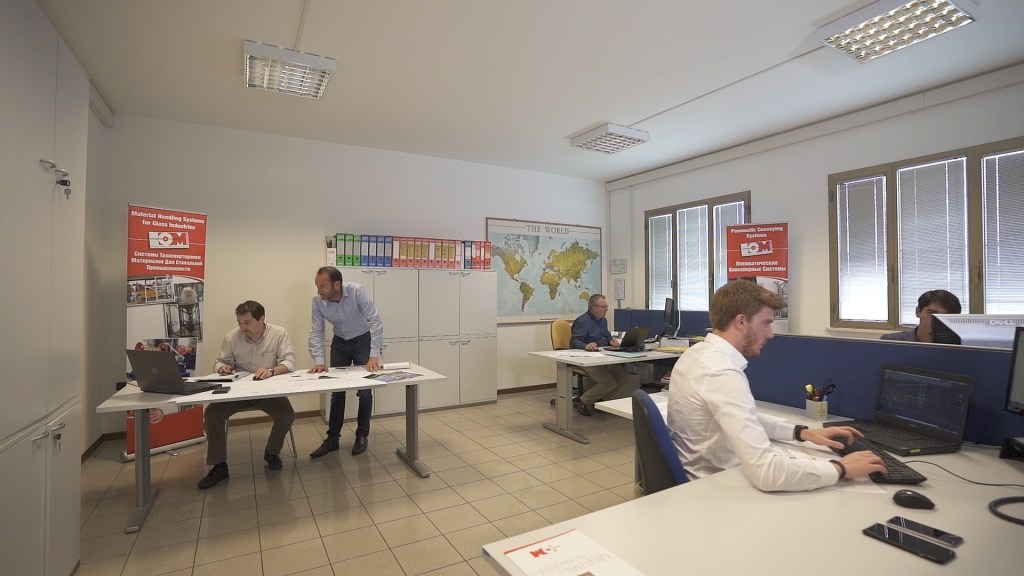 An office scene at OM Siderurgica showing employees working on computers and collaborating over plans, with a world map and posters in the background.