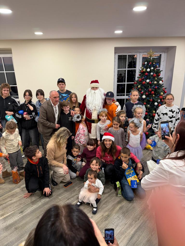 A group of children and adults, including Santa Claus, gathered for a festive celebration at the Cascina Torta Reception Center in Turin, decorated for the holidays.