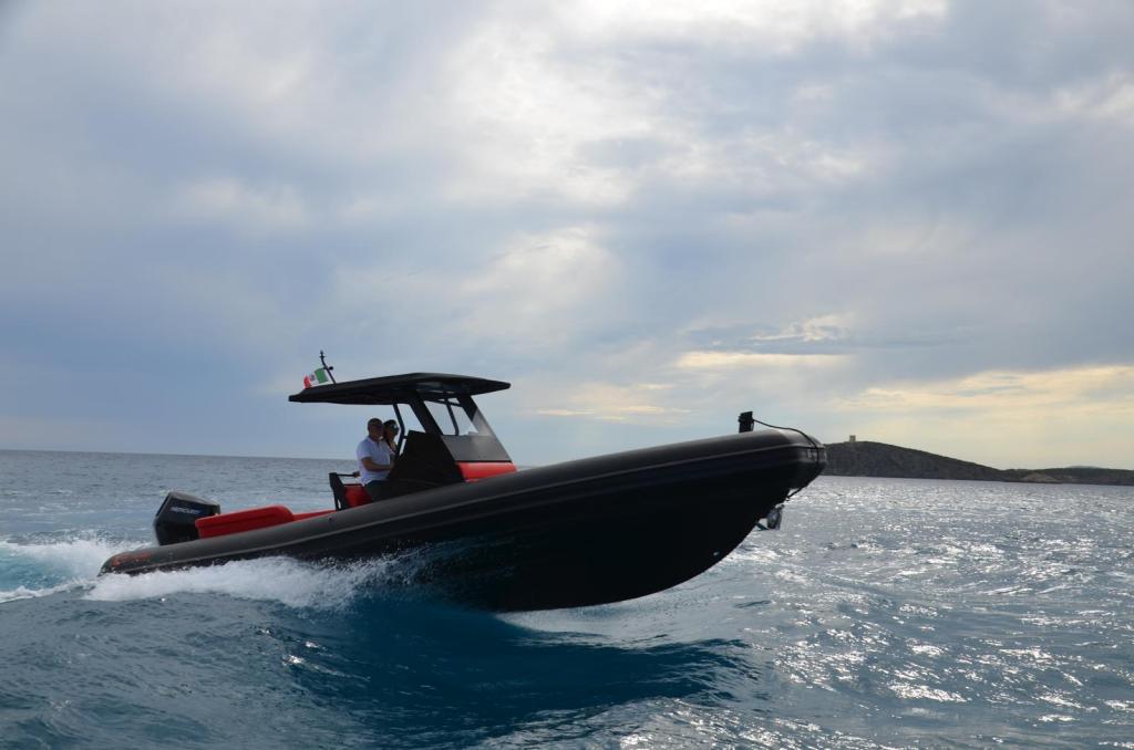 A sleek black boat cruising through the turquoise waters off the coast of Sardinia, with two individuals aboard, one at the helm. The scene captures an expansive sky with clouds, and a distant island on the horizon.