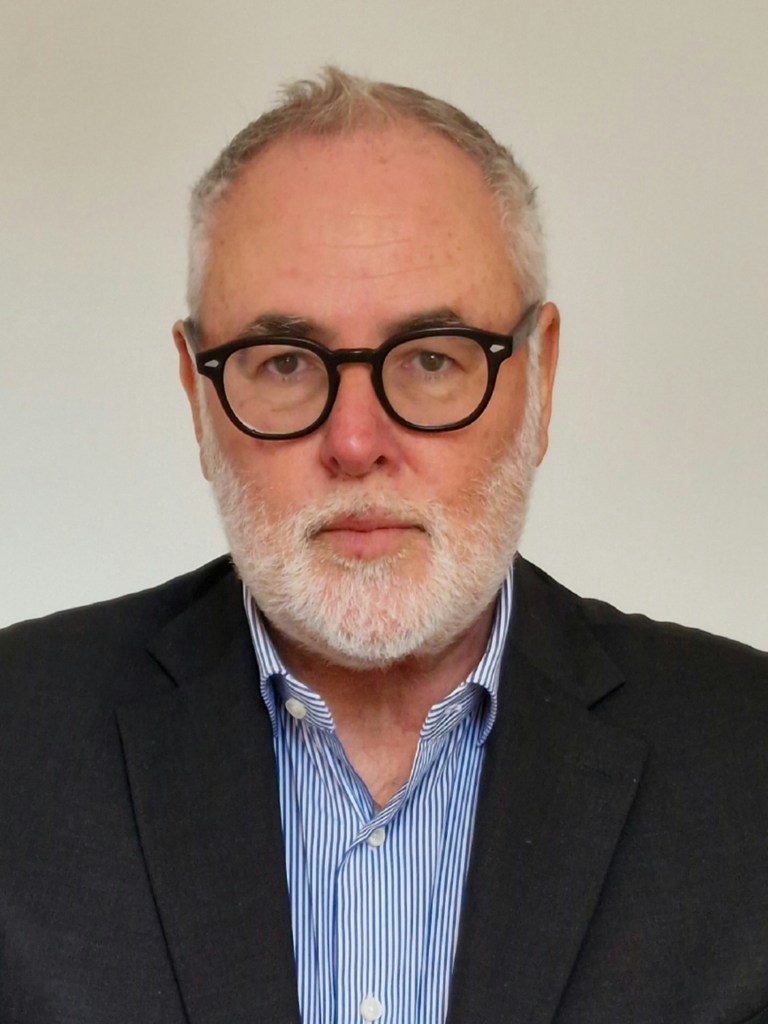 Portrait of a man with glasses and a beard, wearing a dark blazer over a striped shirt, posing in front of a neutral background.