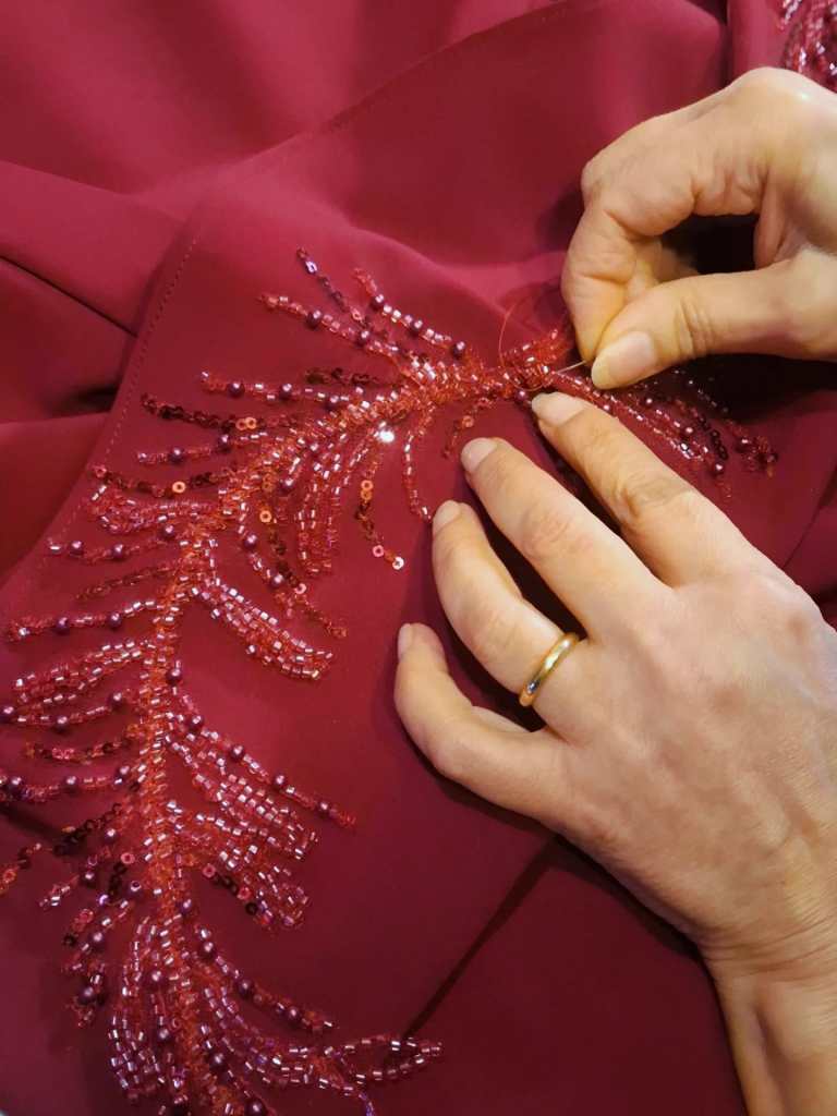 A close-up of hands sewing intricate red beaded embroidery onto a burgundy fabric.