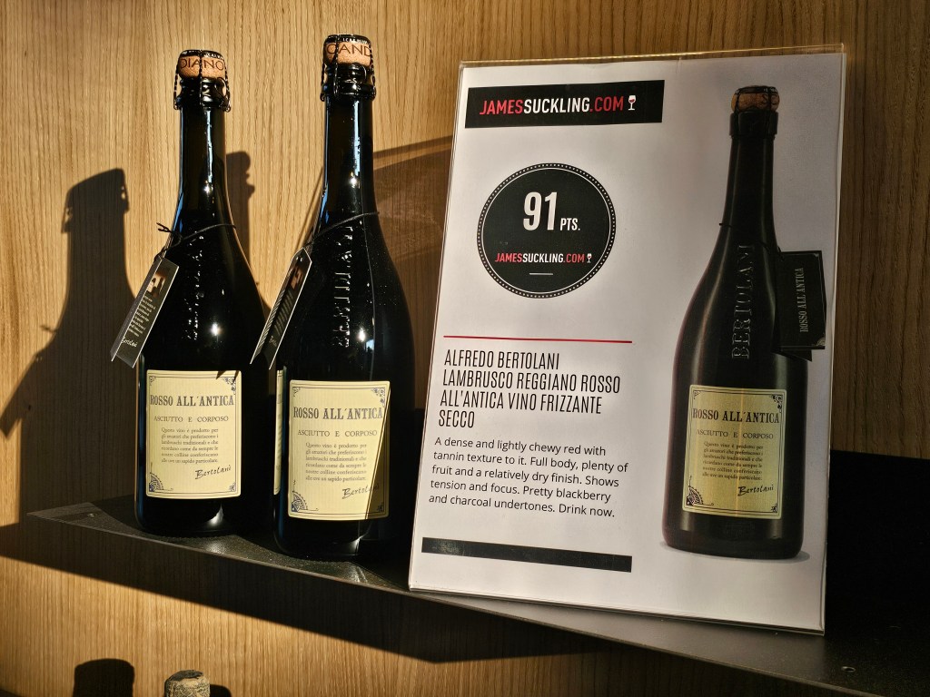Two bottles of Rosso All'Antica wine by Alfredo Bertolani displayed on a wooden shelf, accompanied by a tasting note and rating poster highlighting its flavor profile and characteristics.