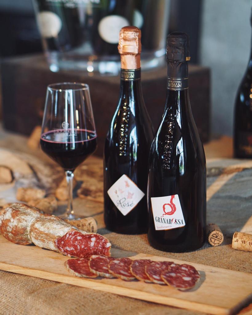 A wooden board featuring sliced salami, accompanied by two bottles of wine, one labeled Rosé and the other Granarossa, along with a glass of red wine in the background.