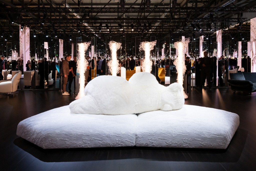 A large, plush, white sofa shaped like a sleeping figure, surrounded by a crowd of people in an exhibition space with striking lighting and mirrors.