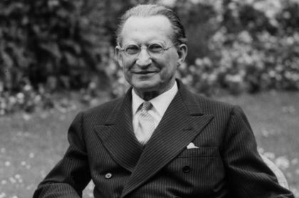 A black and white portrait of an elegantly dressed older man with glasses, smiling while seated outdoors in a garden.