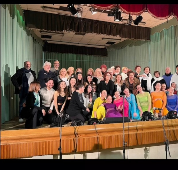 A large group of performers posing together on a stage, wearing a variety of colorful outfits, with a backdrop of green curtains.