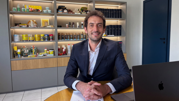 A man in a suit sitting at a desk with a laptop, smiling, in an office setting with shelves displaying various products and items.