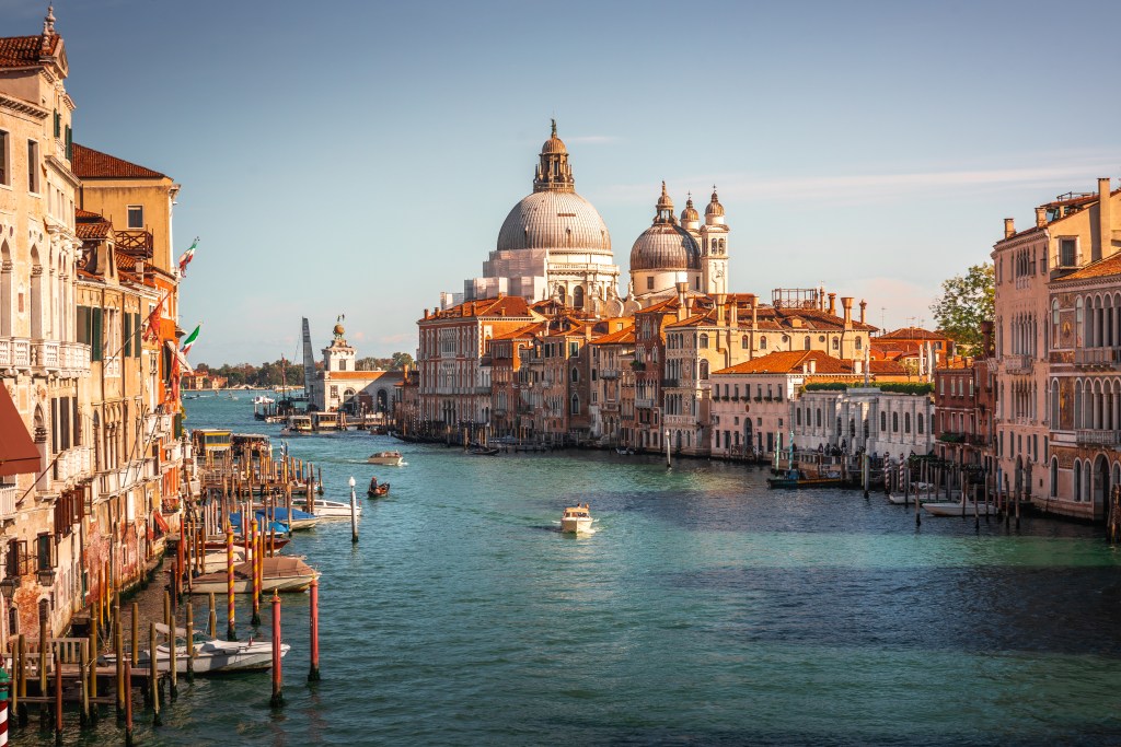 A scenic view of the Grand Canal in Venice, featuring historic buildings and boats, with a large dome structure in the background under a clear blue sky.