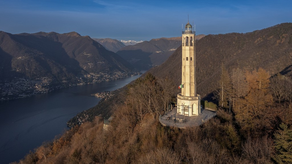 A tall tower with a clock and a lantern situated on a hilltop overlooking a lake surrounded by mountains.