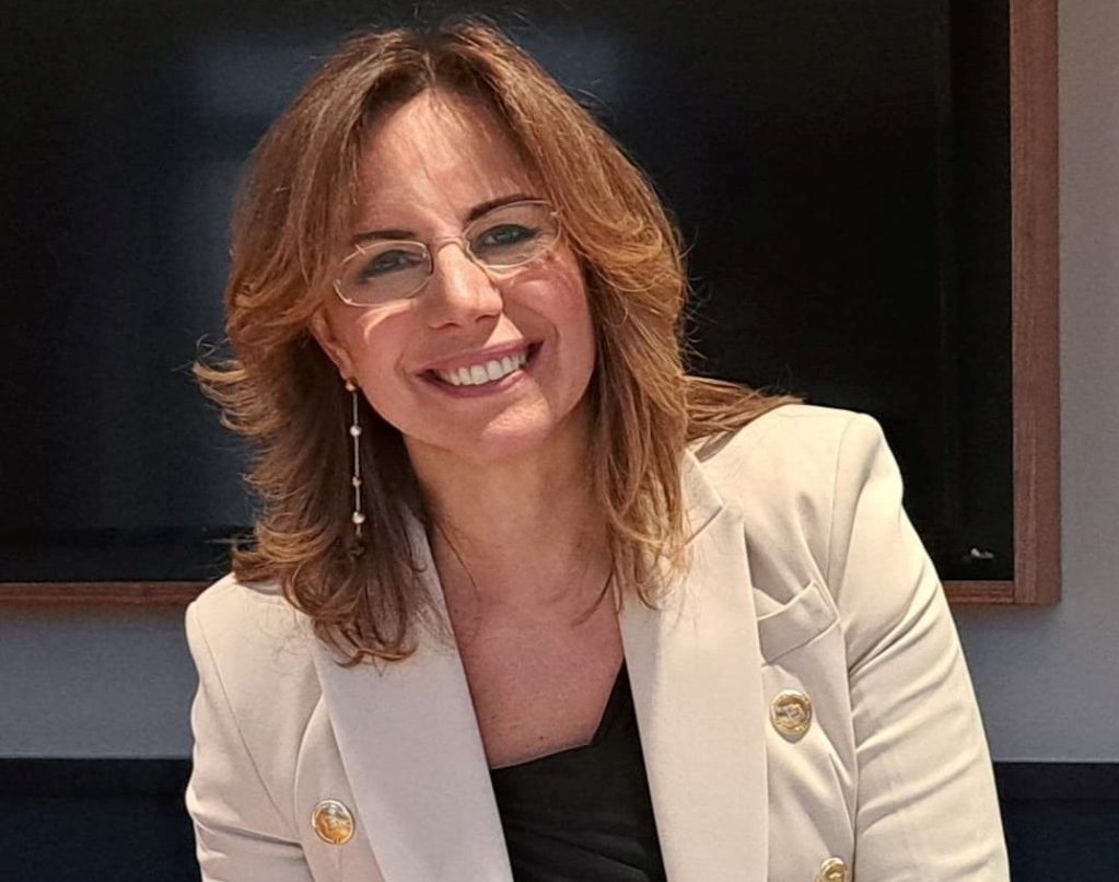 A woman with shoulder-length, wavy hair smiles at the camera while wearing glasses and a light-colored blazer. She appears friendly and approachable in a professional setting.