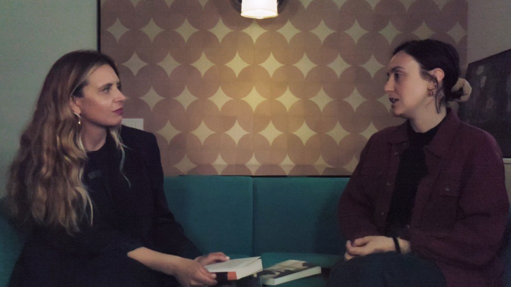 Two women engaged in a conversation while seated on a turquoise couch, with a patterned wall and a lamp in the background. One woman holds a book.