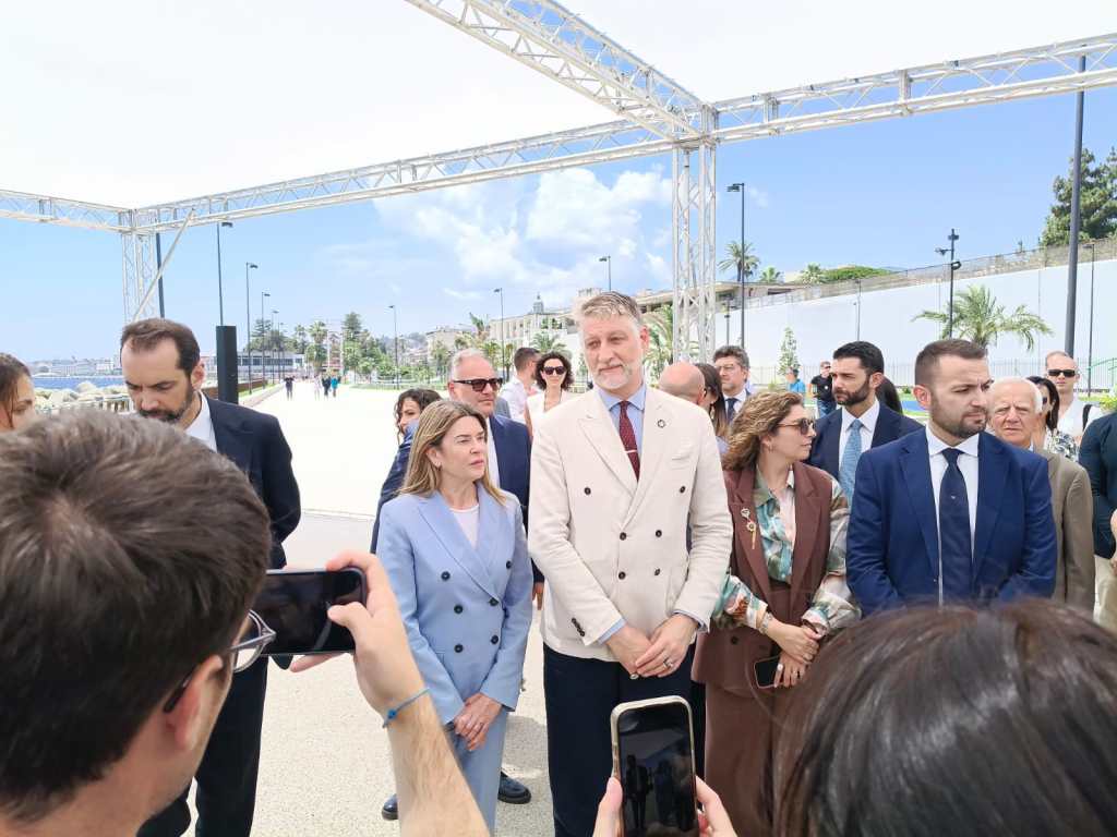 A group of people gathered outdoors under a canopy, engaged in conversation, with a waterfront and palm trees in the background.