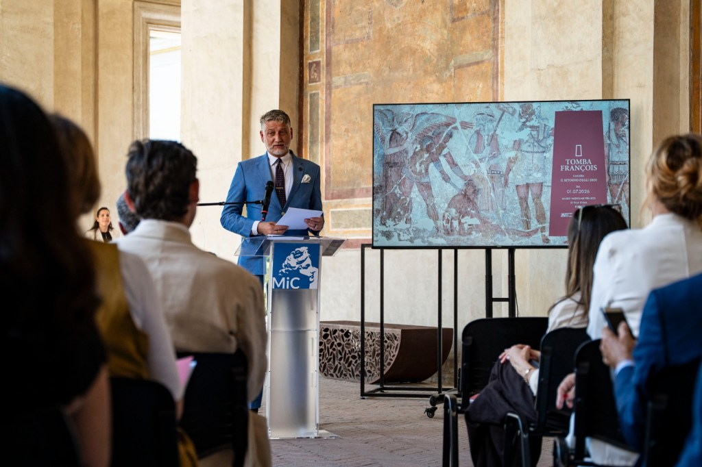 A speaker presents at an event in an architectural setting, beside a screen displaying an artwork titled 'Tomba François.' The audience is seated, listening attentively.