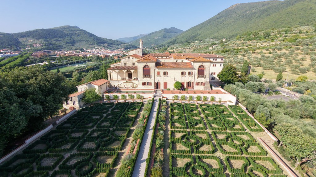 Aerial view of a historic villa surrounded by scenic mountains and gardens with neatly trimmed hedges in intricate patterns.