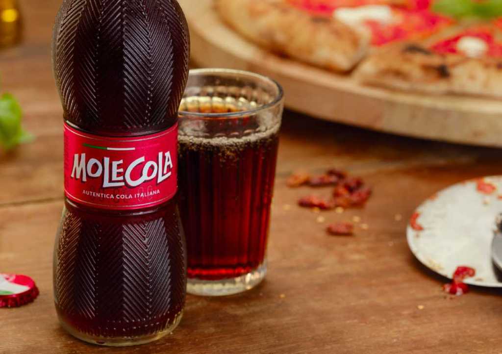 A bottle of Molecola, an Italian cola, next to a glass filled with the drink, set on a wooden table with pizza in the background.