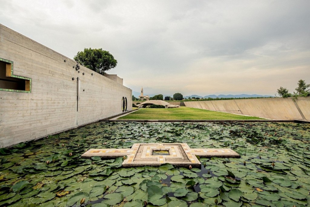 A serene landscape featuring a still pond filled with lily pads, set against a backdrop of modern concrete walls and lush green grass under a cloudy sky.