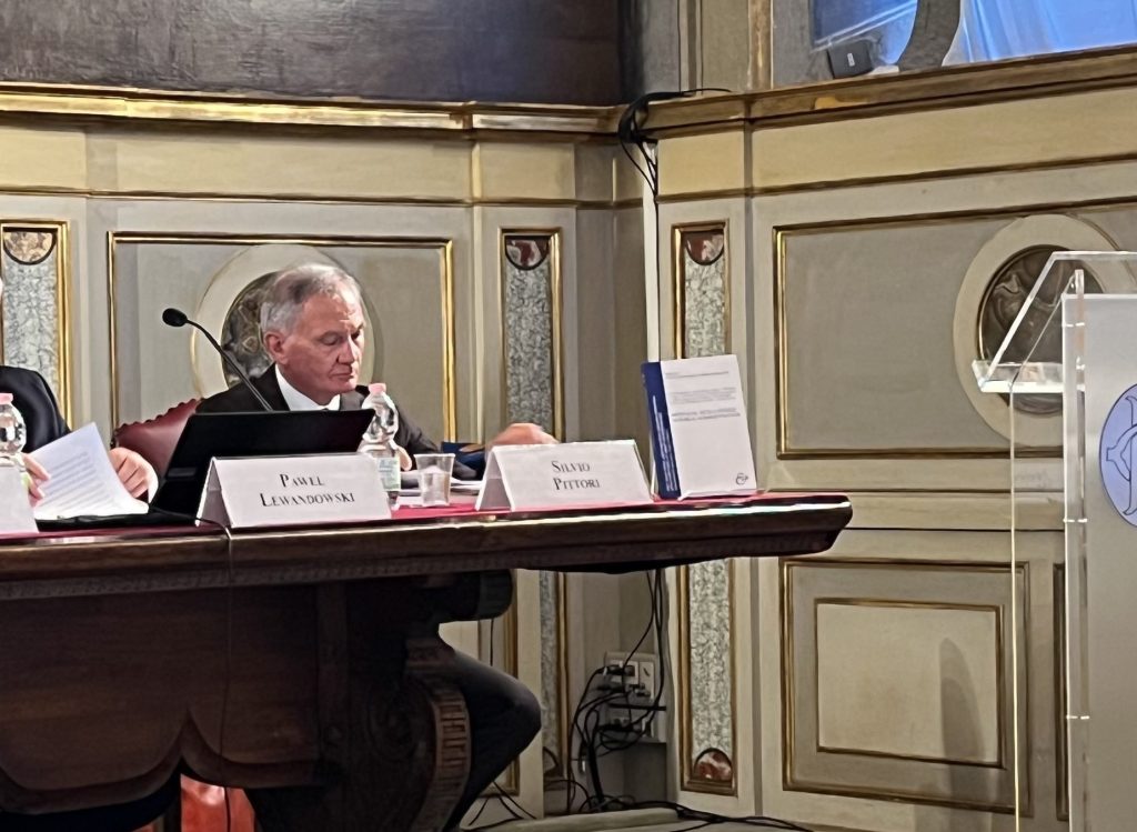 A panel discussion taking place in an ornate room, featuring two speakers at a table with papers and a microphone, while a podium with a logo is positioned on the right.