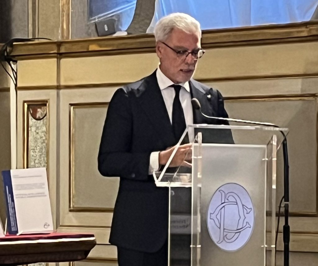 A man in a black suit and tie stands behind a transparent podium, delivering a speech at a formal event.