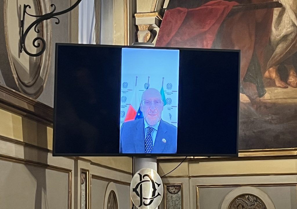 A close-up of a video conference screen displaying a middle-aged man in a suit against a backdrop of flags and emblems, set in a formal interior with decorative walls.