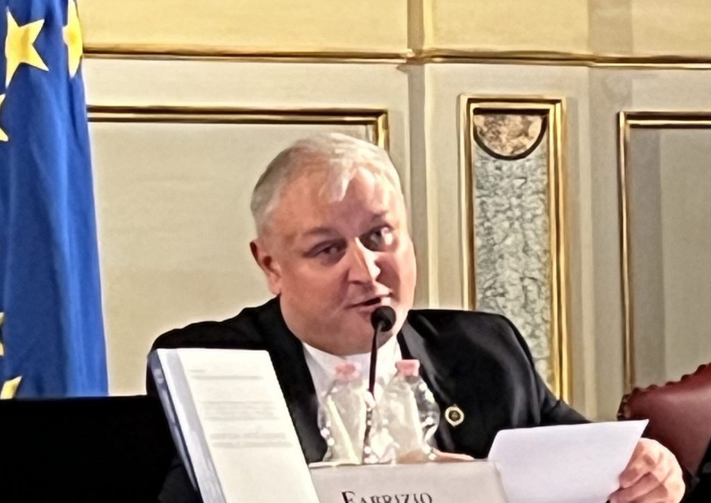 A man speaking at a panel, seated at a table with papers and a microphone, in front of a European Union flag.