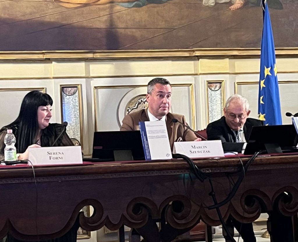 Three individuals sitting at a conference table, with a European flag in the background. The woman on the left has long dark hair, the man in the center is speaking, and an older gentleman is seated on the right, looking towards the speaker.