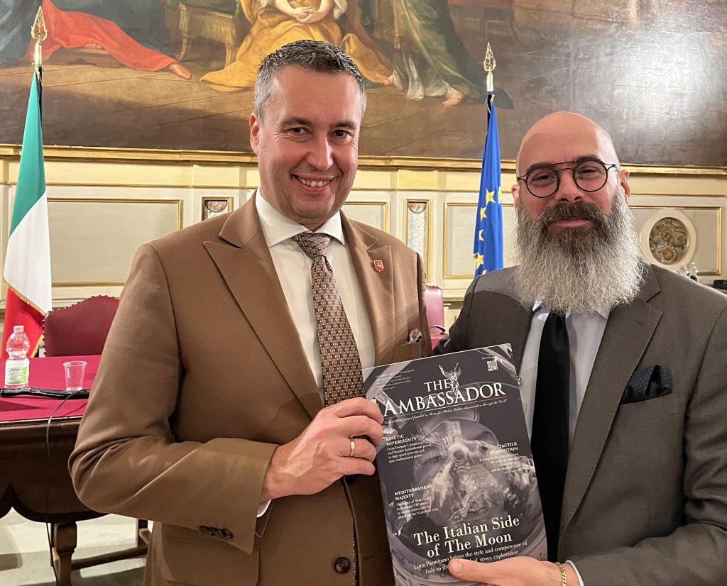 Two men smiling and posing together, one holding a magazine titled 'The Ambassador: The Italian Side of The Moon'. They are in a formal setting with a flag of Italy and the European Union in the background.