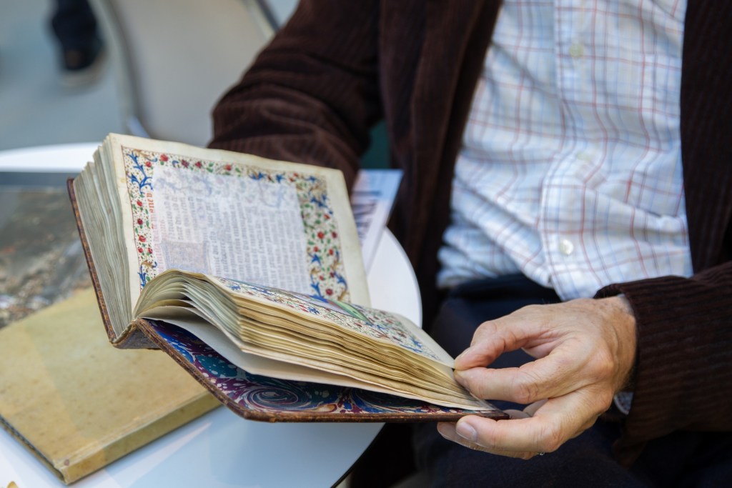 An individual holding an open, ornate book with illustrated pages, seated at a table.