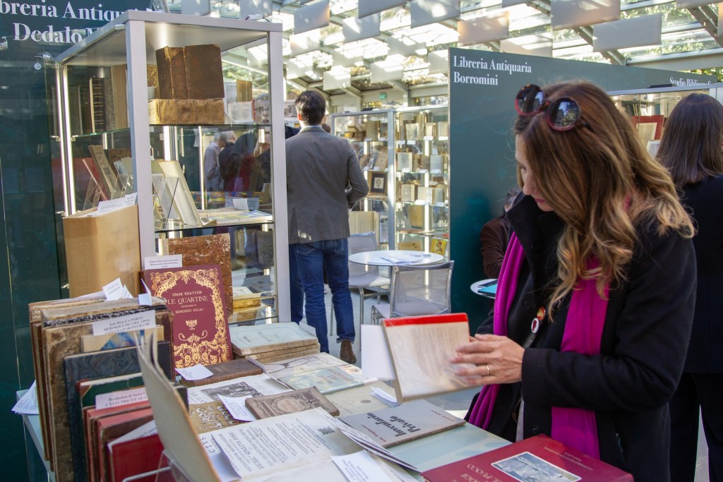 A woman inspecting a book in a vintage bookstore, surrounded by shelves of antique books and other customers browsing.