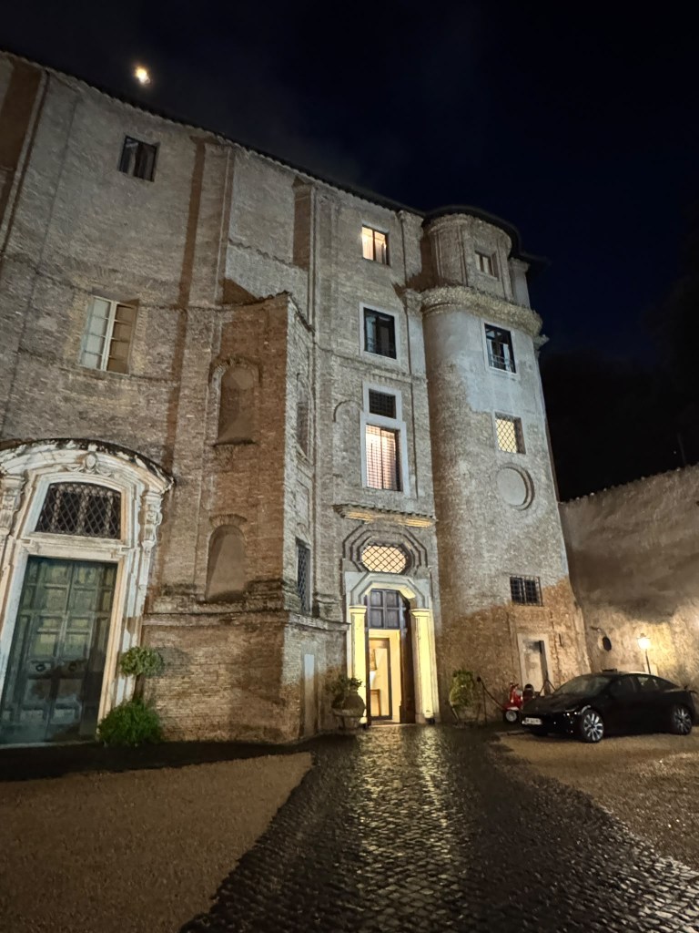 View of a historic building at night, showcasing a brick facade, illuminated door, and parked car in a cobblestone courtyard.